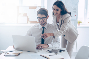 woman pointing at laptop with smile and discussing something with her coworker while standing at office