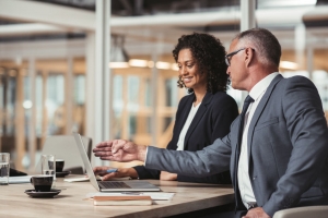 Mature businessman and young work colleague talking together and working on a laptop while sitting at a table in an office boardroom